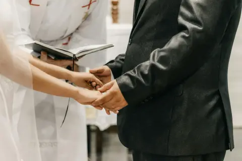 Mani che si stringono durante la dichiarazione matrimonio, con un libro aperto e un sacerdote sullo sfondo.
