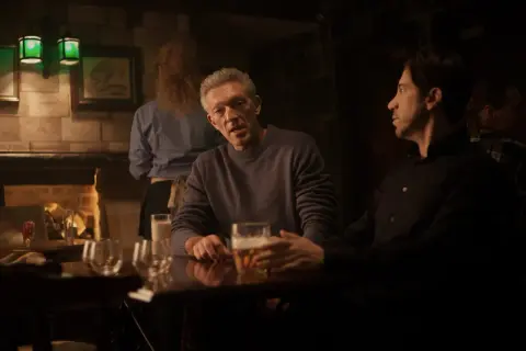 Besetzung von Damaged: Vincent Cassel und Reda Kateb sitzen in einer dunklen Bar bei Bier.