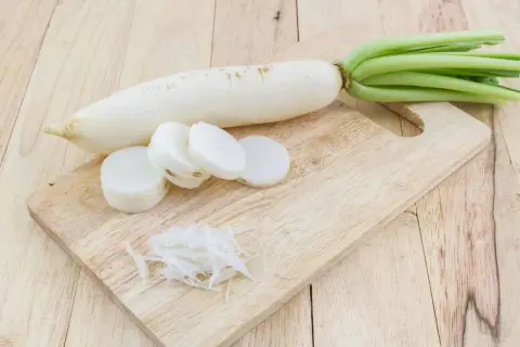 Ein weißer Rettich liegt auf einem Holzbrett, daneben liegen Scheiben und geraspelte Stücke. Perfekt, um Rettich zu fermentieren.