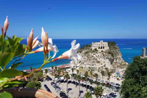Fiori bianchi in primo piano, sullo sfondo il Santuario di Santa Maria dell'Isola a Tropea, un must per chi cerca cosa vedere in Calabria. Spiaggia affollata e mare azzurro.