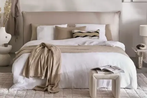 Ein gemütliches Schlafzimmer im shabby stil mit einem weißen Bett, beige Decke und Kissen. Ein Beistelltisch mit Lampe und Deko.
