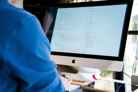 Programista IT w niebieskiej bluzie pochyla się nad monitorem z kodem.