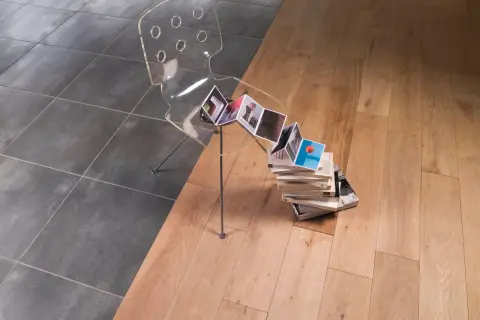 Une barre de seuil discrète sépare un carrelage sombre d'un parquet clair. Une chaise transparente et des livres créent une scène artistique.