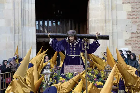 Imagen de una **procesión Miércoles Santo**. Una figura de Jesús carga la cruz, rodeada de nazarenos con túnicas moradas y capirotes dorados.