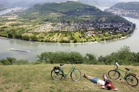 Pareja descansa junto a sus bicicletas en la ruta EuroVelo 15, con un río y un pueblo en el valle.