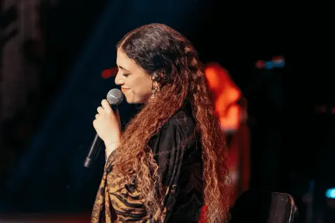 Cantante con lunghi capelli ricci, microfono in mano, sul palco. Un'espressione che evoca la bellezza della Sicilia, un testo che risuona.