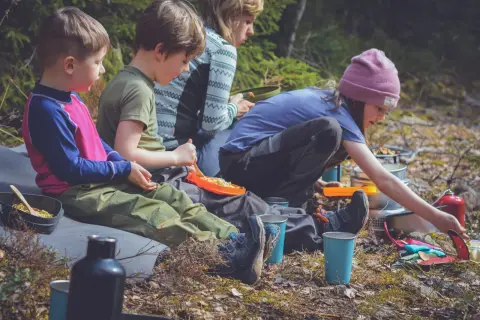 Familj njuter av en picknick i skogen, kanske vid restaurang Ånnaboda. Barn äter pasta och vuxna förbereder mat.