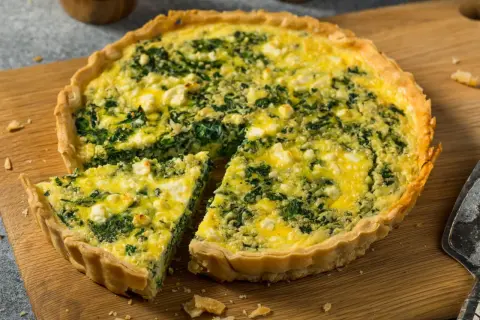 Pyszna tarta ze szpinakiem i serem feta, z jednym kawałkiem już wyciętym, prezentuje się apetycznie na drewnianej desce.