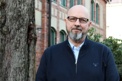 Kaj Stenman, en man med helskägg och glasögon, står framför en tegelbyggnad.