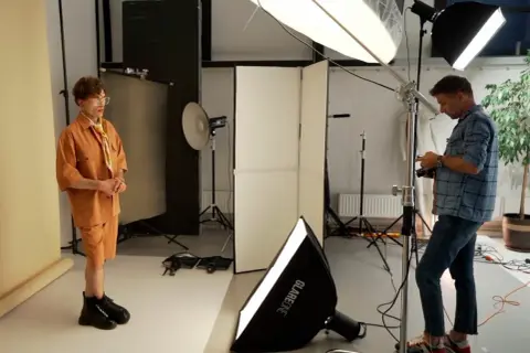 Model w pomarańczowym stroju pozuje w studiu, gdzie fotograf ustawia sprzęt, tworząc idealne naturalne źródła światła.