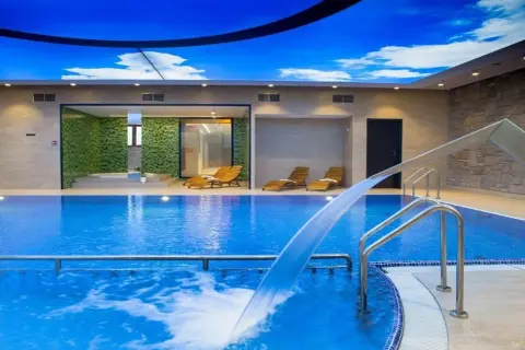 Relaks w basenie hotelu Crocus pod błękitnym niebem. Woda kaskadą spływa do basenu, obok leżaki i zielone ściany.