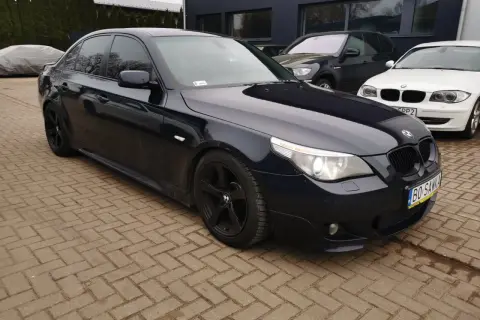 Ciemnogranatowe BMW E60 535d na czarnych felgach, zaparkowane na kostce brukowej przed budynkiem.