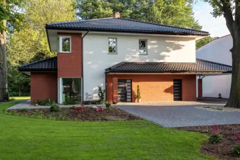 Nowoczesny dom z ceglanymi elementami i dachem kopertowym, otoczony zielenią.