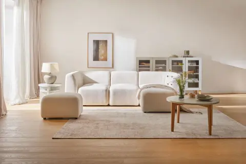 Przytulny salon z kanapą i pufą w odcieniach beżu farby. Jasne zasłony, drewniana podłoga i subtelne dekoracje tworzą spokojną atmosferę.