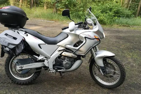 Srebrny motocykl Aprilia Pegaso 650 z kufrem i sakwami, gotowy na przygodę w lesie.
