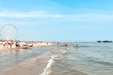 Słoneczny dzień na plaży w Rimini. Ludzie spacerują, bawią się w wodzie, a w oddali widać diabelski młyn. Idealne miejsce na wakacje!