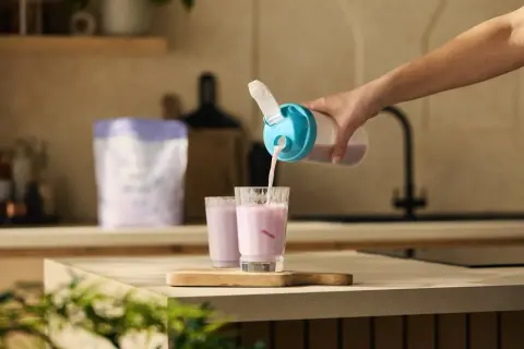 Eine Hand gießt einen rosa Proteinshake aus einem Shaker in ein Glas. So einfach ist es, was bewirken proteinshakes für die schnelle Nährstoffversorgung.