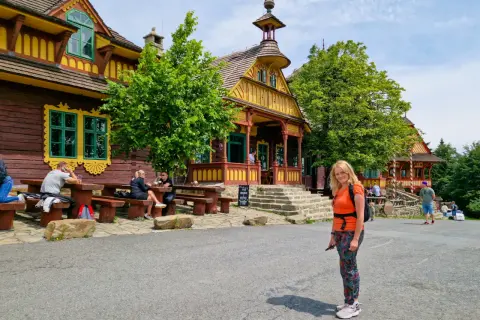 Kolorowe, drewniane budynki w Czechach, pełne ciekawostek dla dzieci. Kobieta w pomarańczowej bluzce pozuje na tle malowniczej architektury.