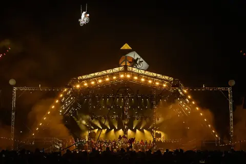 A performer on a bicycle is suspended in the air above a brightly lit stage at Glastonbury. The crowd below anticipates the next act, checking their Glastonbury set times.