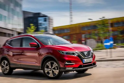 Czerwony Nissan Qashqai 1 jedzie ulicą w mieście. W tle nowoczesne budynki i niebieski znak drogowy.