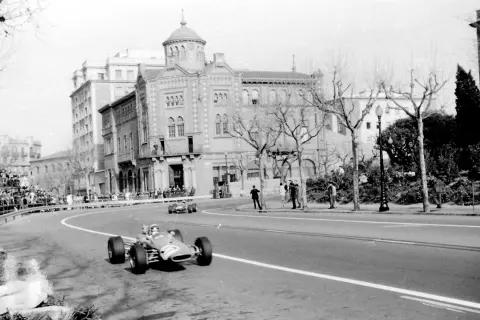 Coches de carreras compiten en el circuito de Montjuïc, con espectadores en las gradas y edificios históricos al fondo.