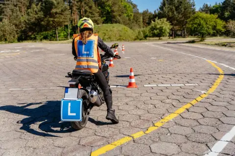 Osoba na motocyklu z tablicą "L" i kamizelką odblaskową na placu manewrowym.