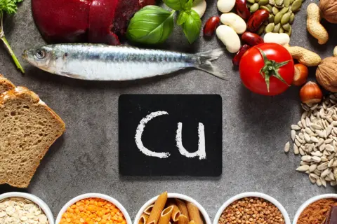 Na stole leżą ryba, chleb, pomidor, orzechy, nasiona i inne produkty bogate w miedź. Tabliczka z napisem "Cu" symbolizuje ten pierwiastek.