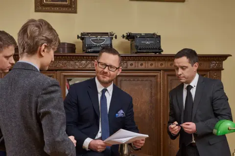 Czterech mężczyzn w garniturach w gabinecie, dyskutujących nad dokumentami.