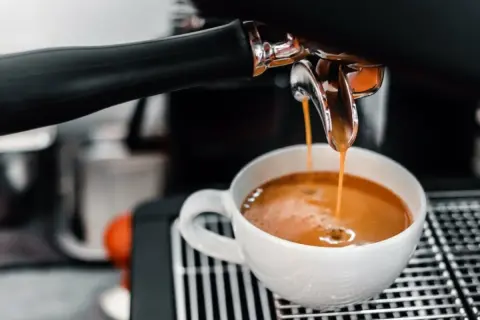 En espressomaskin hjälper till att brygga espresso. Varm, gyllene kaffe rinner ner i en vit kopp.
