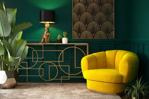 Elegancka tkanina art deco zdobi ścianę nad złotą konsolą z lampą w kształcie kota. Obok żółty fotel i bujna roślina.