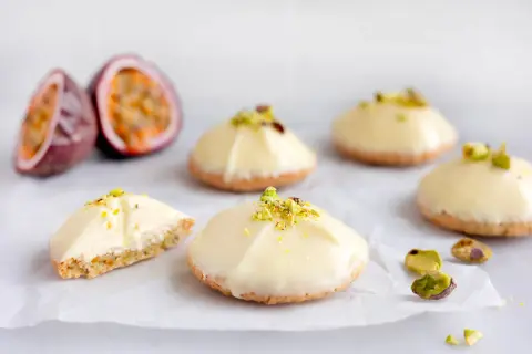 Små, vita bakelser med pistagecrunch, inspirerade av passionsfrukt recept. En är uppskuren och visar en krämig fyllning.