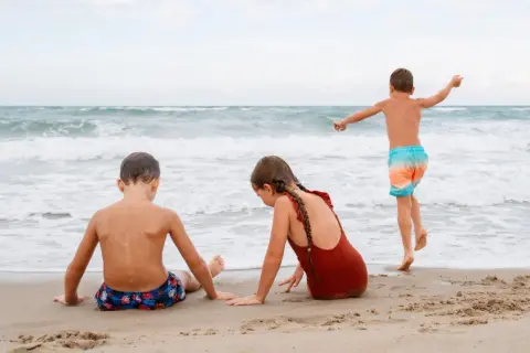 Dzieci bawią się na plaży, szukając **jastarnia atrakcje dla dzieci**. Chłopiec biega w stronę morza, dziewczynka i drugi chłopiec siedzą na piasku.