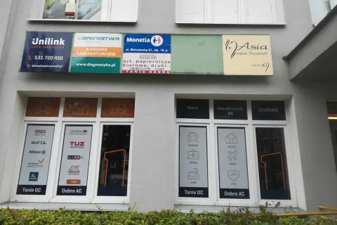 Szyldy firm ubezpieczeniowych i salonu fryzjerskiego na szarej ścianie budynku.