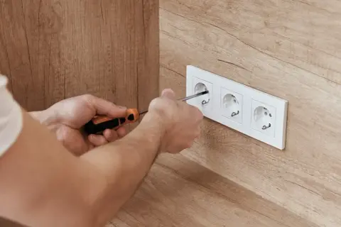 Ręce dokręcają śrubokrętem gniazdko elektryczne. Widać trzy gniazda, a ich rozmieszczenie tworzy pewną **amplitudę** na ścianie.