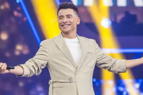 Uśmiechnięty mężczyzna w beżowej marynarce trzyma mikrofon, stoi na tle kolorowej sceny.