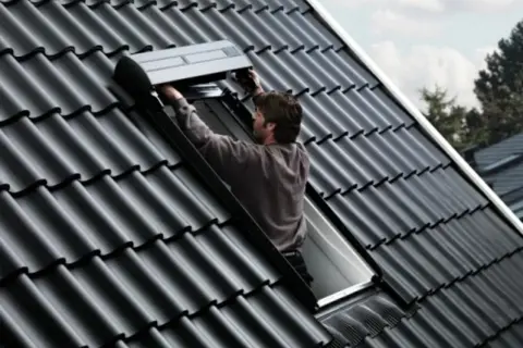 Un couvreur installe un volet roulant sur un toit en tuiles noires, un modèle velux 3uc b01 eu.