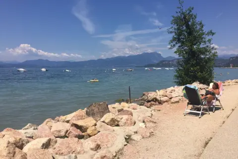 Una donna legge su una sedia a sdraio sulla riva rocciosa di Cisano, Gardasee. Barche galleggiano sull'acqua calma con montagne sullo sfondo.