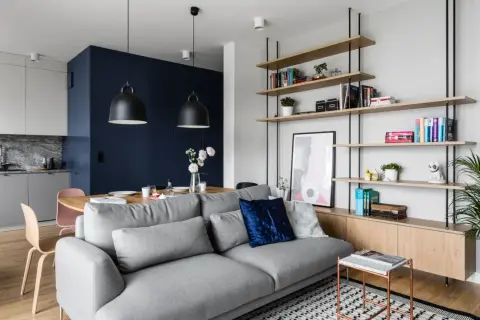 Szara sofa w małym salonie z półkami, stołem i aneksem kuchennym w tle.
