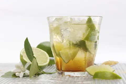 Orzeźwiająca caipirinha przepis: drink z lodem, limonką i cukrem. Obok leżą świeże limonki i kwiatki.