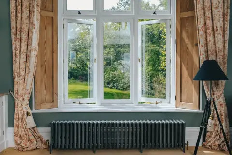 Ventanas blancas abiertas a un jardín verde, con cortinas florales y un radiador antiguo. Las mejores ventanas para disfrutar del exterior.