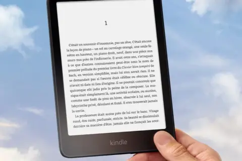 Main tenant un Kindle, un livre numérique de Nextory, sur fond de ciel bleu et nuageux.
