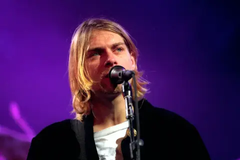 Kurt Cobain śpiewa do mikrofonu na tle fioletowej sceny.