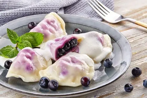 Pyszne pierogi z jagodami, polane śmietaną, ozdobione miętą i świeżymi owocami. Idealne na deser!