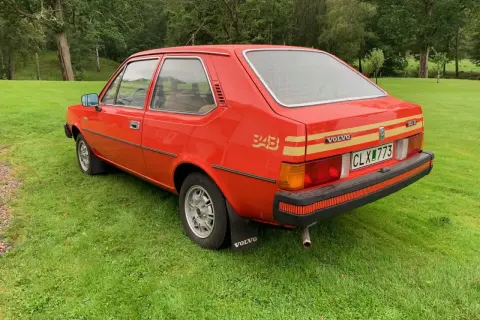 En röd Volvo 343 står på en grön gräsmatta med skog i bakgrunden. Bilen har en gul rand längs sidan och texten "343" i guld.
