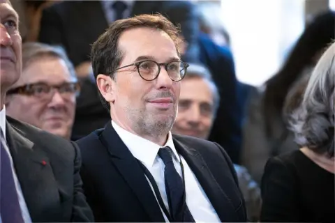 Un homme avec des lunettes, en costume noir, souriant.