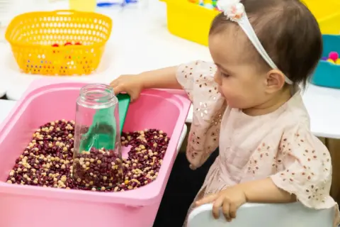 A baby girl explores a winter sensory bin filled with beans and a scoop.