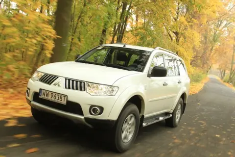 Biały model terenówki Mitsubishi Pajero Sport jedzie po drodze wśród jesiennych drzew.