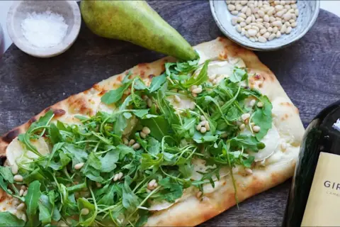 En läcker pinsa med chevre, ruccola och pinjenötter, bredvid en päronhalva, salt och en vinflaska.