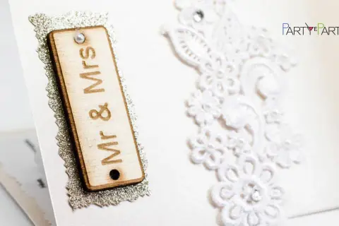 Dettagli di un invito di nozze: targa in legno "Mr & Mrs", pizzo bianco e logo "Party Partenze". Un esempio di diversi tipi di nozze.