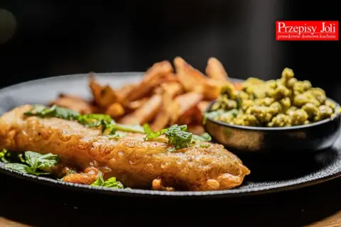 Chrupiące fish and chips z zielonym groszkiem i świeżą miętą na czarnym talerzu.